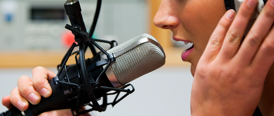 Foto colorida de uma locutora em frente ao microfone. Sua boca está aberta, sua mão direita segura o microfone e a esquerda toca o fone de ouvido. 